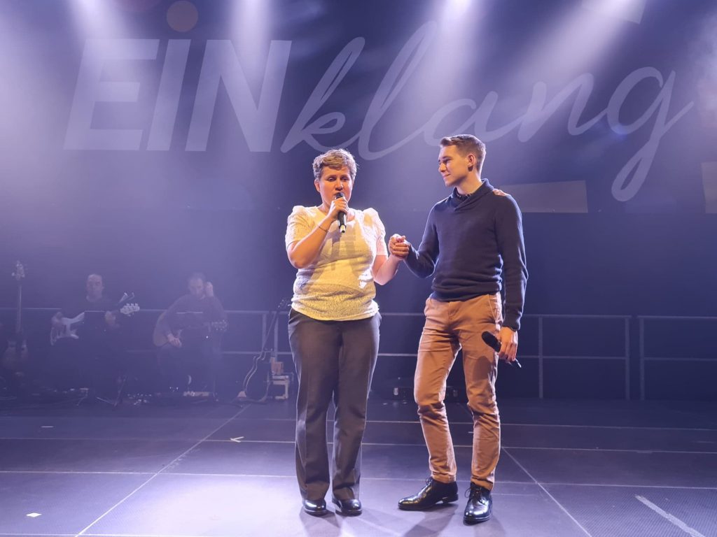 Kobieta i mężczyzna trzymają się za ręce, w dłoniach mają mikrofony, stoją na scenie.