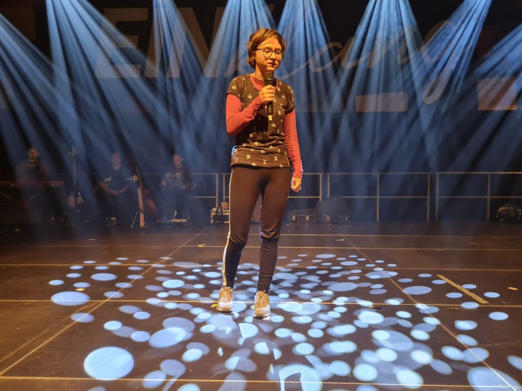 Dziewczynka stoi na scenie oświetlonej scenicznym fleszem, w dłoni trzyma mikrofon.