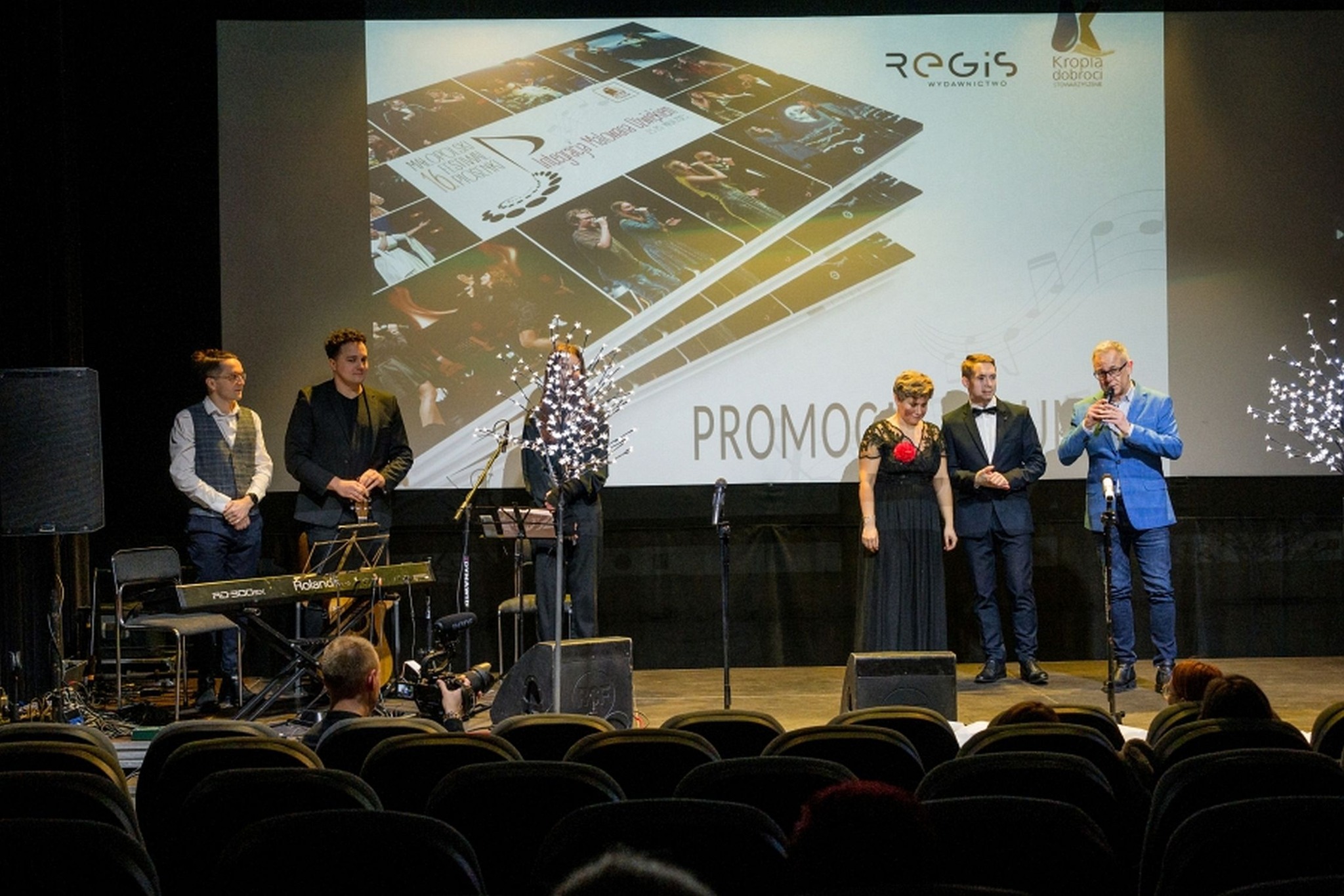 Na scenie stoi kilkoro muzyków i wokalistów, w tle widać okładkę ksiązki.